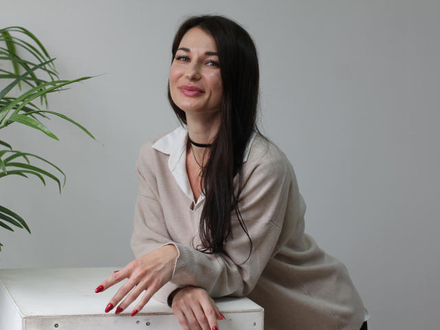 AlexOakman with long black hair, smiling, wearing a beige sweater with a white collar, black choker, and red nails, leaning on a white table with a green plant.
