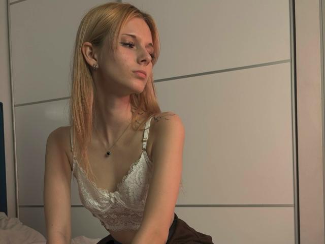 DierdreLorenzano with blonde hair, white lace bra, small tits, and a tattoo on her right shoulder, sitting in a bedroom, looking to the side with a pouty expression.