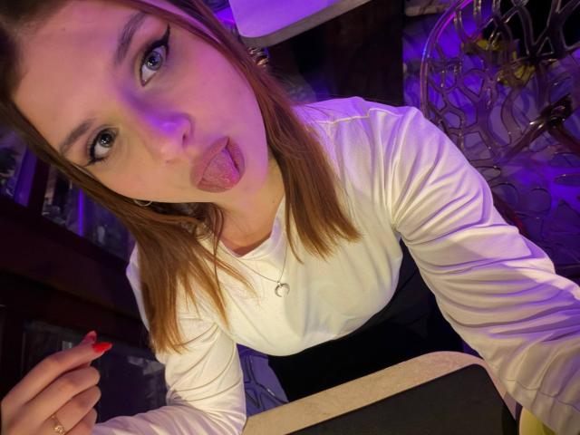 HeikeRippy with straight brown hair, sticking out tongue, blue eyes, white shirt, black skirt, purple lighting, red nails, and a gold necklace, leaning forward, holding something.