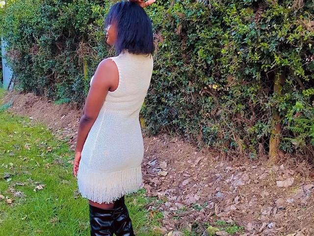 QueenIvy se tient dos à la caméra, vêtue d'une robe blanche en tricot avec des franges et de hautes bottes noires, ses cheveux noirs courts contrastant avec sa peau foncée..