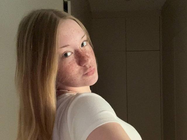 TashinaDufort with freckles, long blonde hair, white tee, looking over shoulder, slight pout, pale skin, subtle makeup, soft lighting, neutral background.