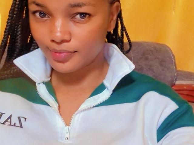 QueenIvy, avec des cheveux tressés, une peau brune et des lèvres roses, portant un sweat à capuche vert et blanc avec fermeture éclair, souriant légèrement, montrant de petites seins mignons et une jolie fesse sur la photo..
