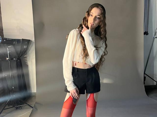 KeraPopham, con cabello castaño largo y ondulado, un top blanco, pantalones cortos negros y medias de gasa rojas, de pie, con una mano en el cuello, fondo gris claro, luces de estudio..