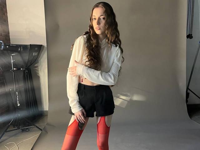 KeraPopham, con cabello largo y castaño, una camiseta blanca de manga larga y corte corto, pantalones cortos negros y medias de gasa rojas, de pie, con los brazos cruzados, en un estudio iluminado por el sol, sosteniendo un teléfono..