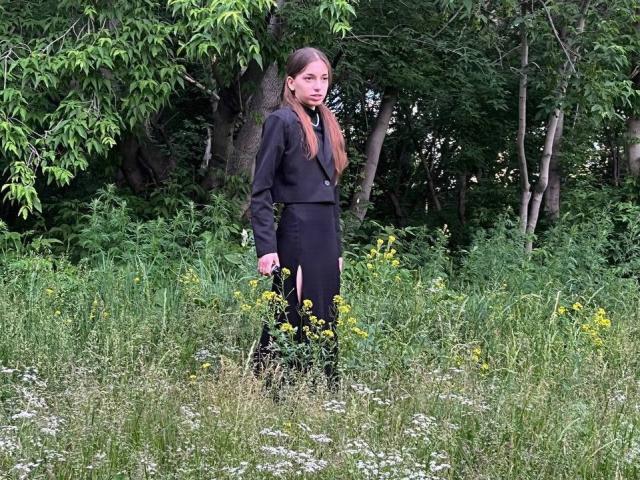 KeraPopham, con cabello castaño, vestida con una chaqueta y falda negros, está de pie en un campo cubierto de hierba, con flores amarillas y trébol blanco, sosteniendo una pequeña bolsa en su mano derecha..