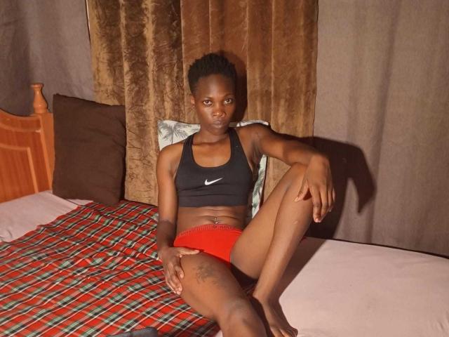MelanyPrincess with dark skin, small boobs, and red panties, sitting on a bed with a plaid blanket, wearing a black Nike sports bra, arm tattoo visible, brown curtains.