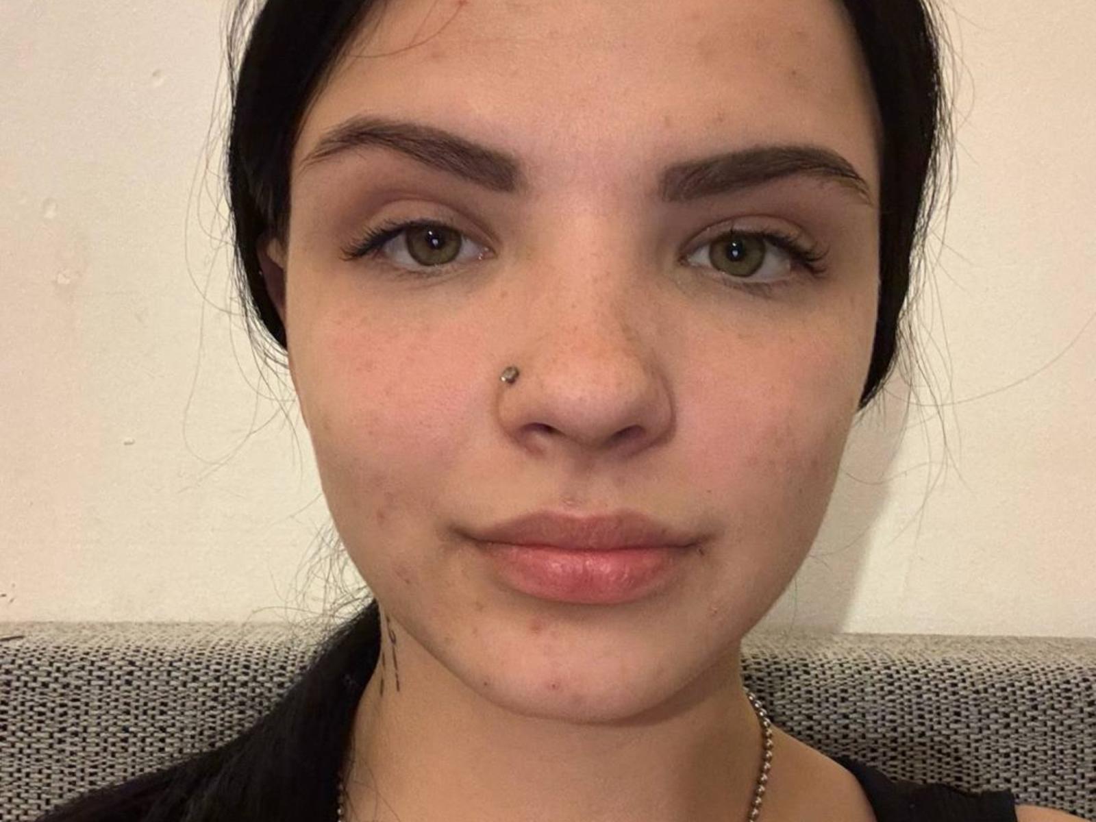 SunshineModert has a cute face with brown eyes, dark eyebrows, a small nose piercing, and pinkish lips. She's wearing a black shirt with a gray textured background.