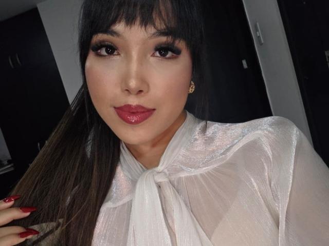 Heyly with shiny white blouse, red lipstick, and long brown hair, holding her hair with red nails, has cute earrings and a slightly smiling face.