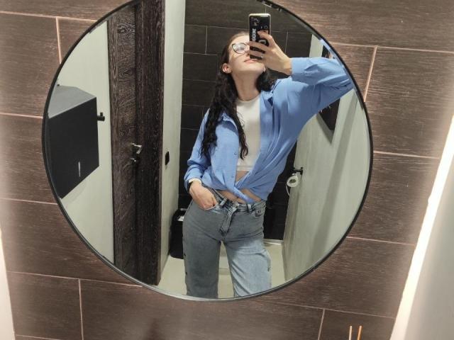NatoshaBurdine with glasses, tied blue shirt, white crop top, and high-waisted jeans, taking a mirror selfie, hand in pocket, head tilted back, long black hair, in a bathroom.