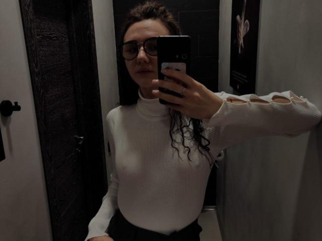 NatoshaBurdine with curly brown hair, wearing glasses, white ribbed long-sleeve top, black skirt, holding phone, mirror selfie, showing small tits, dark door in background.