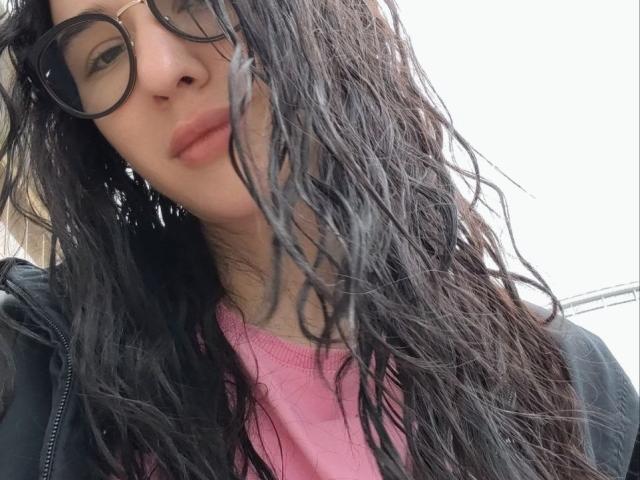 NatoshaBurdine with wet black hair, wearing glasses and a pink shirt, black jacket, slightly parted lips, and a neutral expression, looking down at the camera.