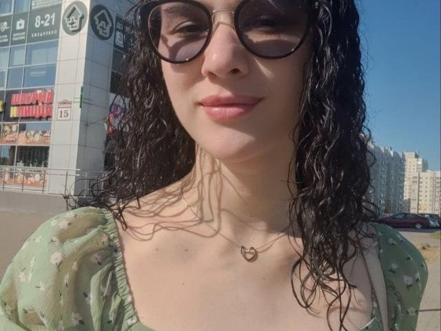 NatoshaBurdine with wet black curly hair, wearing green puffed-sleeve dress with white flowers, black sunglasses, and heart necklace, smiling with pink lips, clear blue sky.