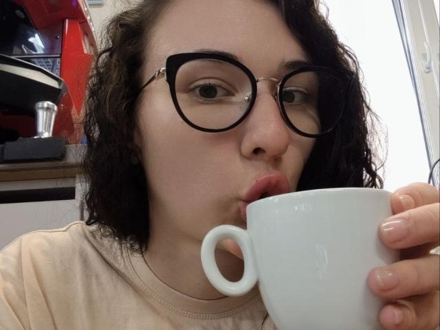 NatoshaBurdine with curly dark hair, black glasses, and pink lips, sipping from a white ceramic mug, wearing a beige shirt. Her hand holds the mug near her mouth.