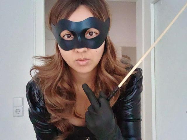 HerrinKira-hot with brown wavy hair, black mask, black leather top, black gloves, holding a wooden stick, making a 'shhh' gesture with fingers.