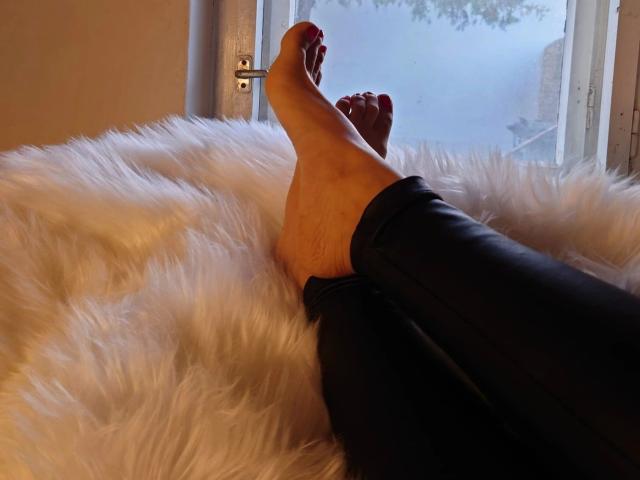 HerrinKira-hot's bare feet with red-painted toenails, crossed at the ankles, resting on a fluffy white rug, wearing black pants, near a window.