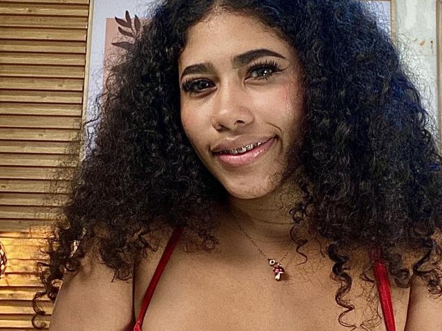 EmiStar, con cabello negro rizado, piel morena y una sonrisa que muestra dientes de metal, vestida con una blusa roja y un collar con un pequeño dije, mirando a la cámara..
