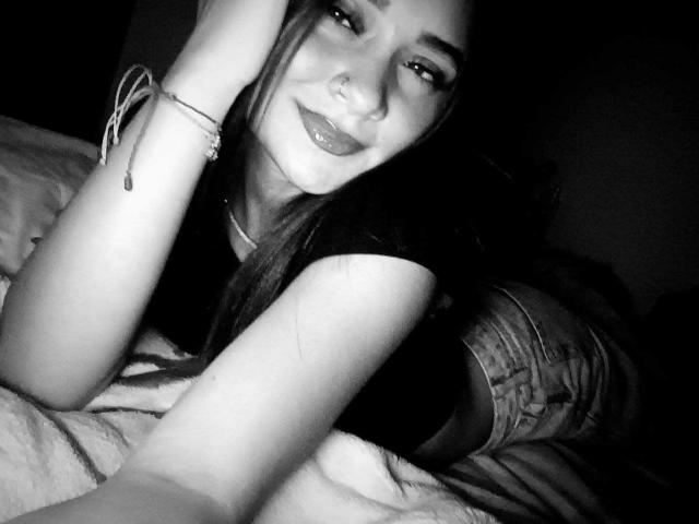 MilaDreams, with long dark hair, nose ring, and black shirt, lays on bed, smiling, with hand on head, wearing denim shorts, in black-and-white selfie.