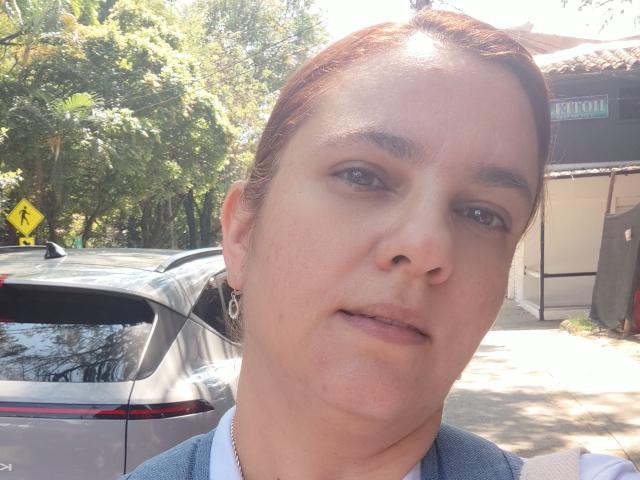 AmeliaWilson has a slight smile, brown eyes, and light skin with subtle freckles. She's wearing a blue top and silver earrings, with a silver car behind her.