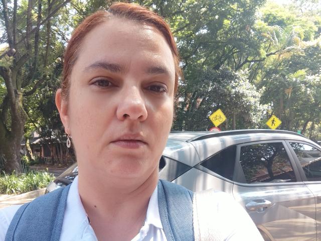 AmeliaWilson with red hair, blue vest, white shirt, and serious expression in front of a silver car with yellow road signs. Her ass is not visible.