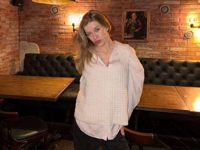 DeedraGalarneau with blonde hair, white textured blouse, and black pants, standing in a dimly lit brick-walled room, leaning slightly with a relaxed, confident expression.