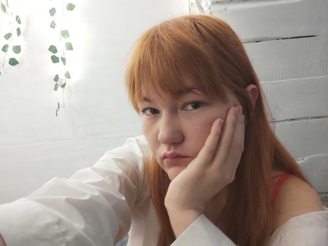 SheritaChieng has red hair, fair skin, and is resting her hand on her cheek with a pouty expression. She's wearing a white shirt with red straps.