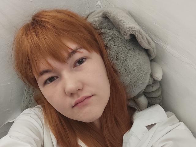 SheritaChieng with red hair, white shirt, and soft gray stuffed animal behind her head, looking slightly down with a neutral expression. Her small nose and light skin are visible.