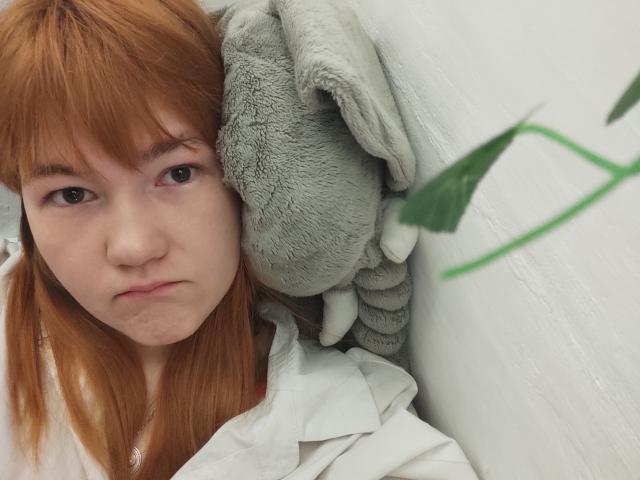 SheritaChieng with red hair, pouty expression, holding gray stuffed animal, white shirt, green leaf in foreground, looking slightly annoyed, soft skin, light makeup, bangs covering part of forehead.