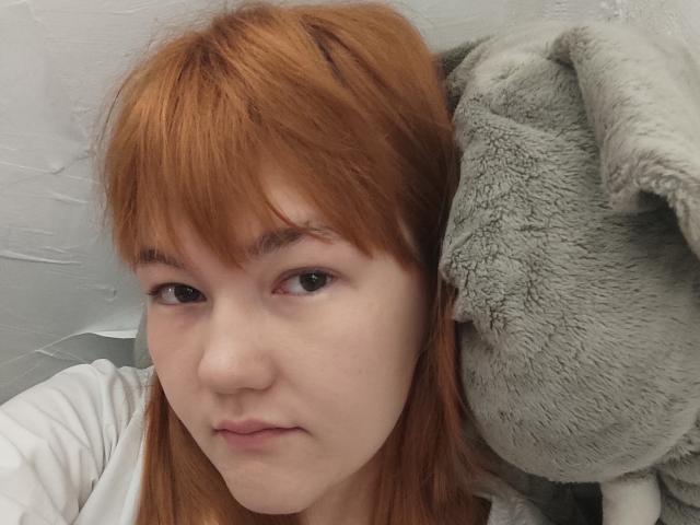 SheritaChieng has red hair, fair skin, and a neutral expression. She's wearing a white shirt, with a gray, fuzzy stuffed elephant on her right.