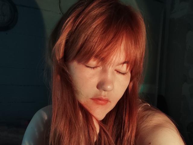 SheritaChieng with red hair, eyes closed, soft sunlight on her face, pale skin, slight pout, bare shoulders, gentle shadow on her right side.