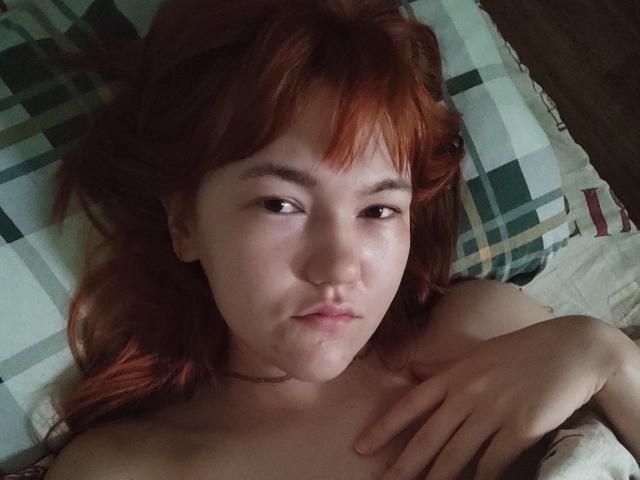 SheritaChieng with red hair, pale skin, and a pouty face, lying on a plaid pillow, topless, hand on chest, choker around neck, slight wetness on her lower lip.