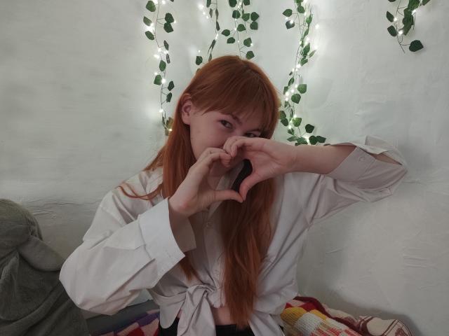 SheritaChieng with red hair, wearing a white shirt, makes a heart shape with her hands in front of her face, sitting on a bed with colorful blankets.