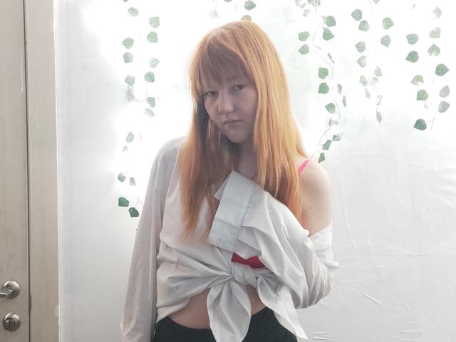 SheritaChieng with red hair, wearing a tied-up white shirt, exposing a pink bra, looking bored, standing in front of a white wall with green leaf decorations.