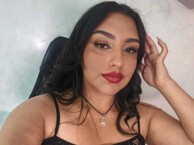 SyraPalmer tem cabelos escuros e ondulados, olhos castanhos, batom vermelho e usa um colar preto com um pingente. Ela está tocando a cabeça com uma mão, mostrando os seios..