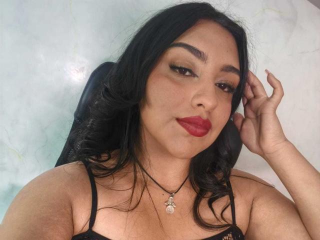 SyraPalmer con capelli neri, rossetto rosso e lingerie nera, indossa una collana, ha una mano tra i capelli, guarda la fotocamera, pelle media, occhi marroni, leggero sorriso, sexy, sicura di sé..