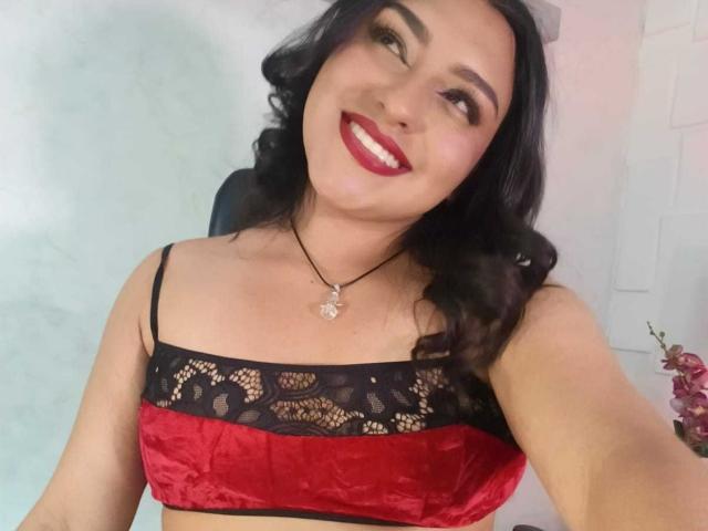 SyraPalmer with black lace and red velvet bra, smiling, dark wavy hair, red lipstick, black strap, necklace with star pendant, holding phone, taking selfie.
