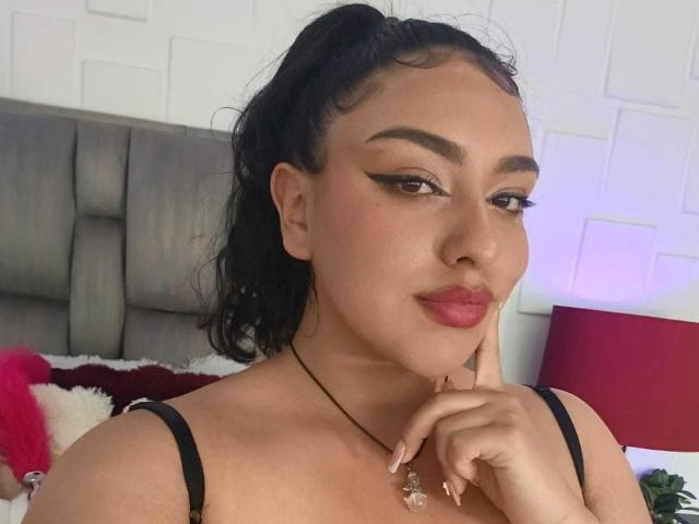 SyraPalmer has dark hair in a ponytail, thick eyebrows, winged eyeliner, pink lipstick, black strap top, and a necklace with a crystal pendant, touching her chin.