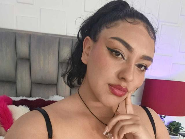 SyraPalmer with dark hair in a ponytail, wearing black straps, has bold eyeliner, pink lipstick, and touches her chin with a finger, wearing a black necklace.