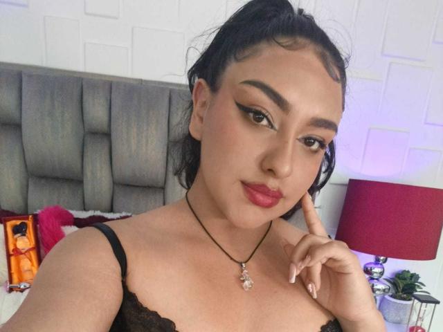SyraPalmer with dark hair in a ponytail, wearing red lipstick, black lace top, and a necklace with a pendant, touching her chin with a finger. Boobs visible.