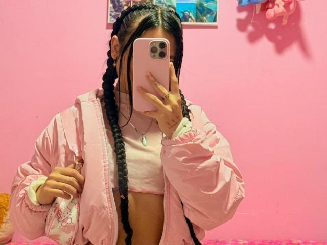 LunaDesire with braids, wearing a pink jacket, pink crop top, and necklace, taking a mirror selfie with her phone, showing her flat stomach and small tits.