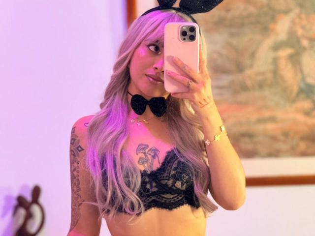 LunaDesire with long, wavy, pinkish-blonde hair, black lace bra, black bunny ears, black bowtie, and tattoos, taking a selfie in a mirror, showing her tits.