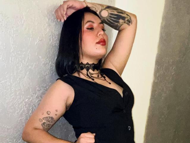AmyRoss with black hair, red lipstick, black choker, black sleeveless top, tattoos on arm and shoulder, hand on wall, standing with arm raised, showing her tits.