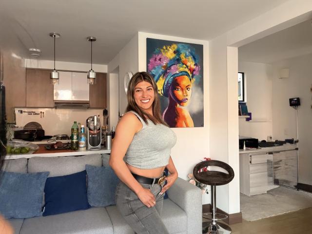 LunaBeya with big boobs, smiling, wearing a gray crop top and jeans, standing in a kitchen, next to a colorful painting of a woman with a colorful headpiece.