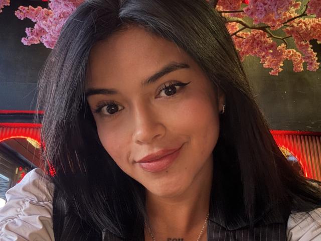 MajoJones has dark, straight hair, brown eyes with winged eyeliner, and a subtle smile. She's wearing a white shirt with puffed sleeves, and a small gold earring.