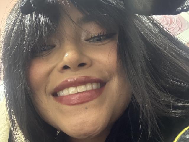 MajoJones has dark, straight hair, one eye closed, smiling with red lipstick, showing white teeth, and a small mole on her lower lip. She's wearing a black hat.