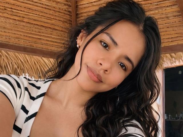 EvaPrincess has long, wavy black hair, brown eyes, and a cute smile. She's wearing a striped top, showing her tits slightly, with a wooden ceiling above.