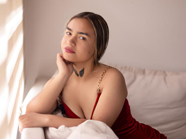 AnnaLadera with brown hair, red velvet dress, gold chain strap, resting chin on hand, looking up, small black tattoo below neck, white pillow, soft sunlight, slight smile.