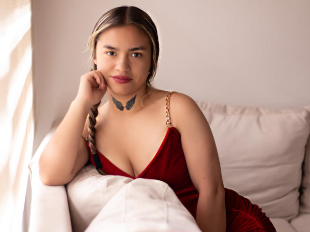 AnnaLadera with light brown skin, red velvet dress, and butterfly necklace, resting on a white couch, looking at the camera with her hand on her chin, showing her tits.