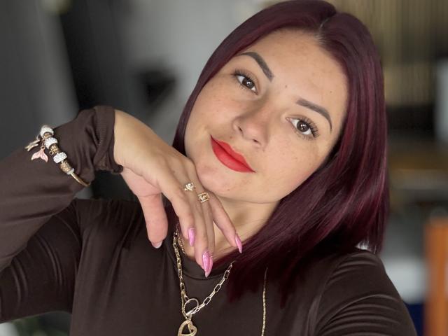 KatherineToro with red lipstick, dark brown long-sleeve shirt, pink nails, red-brown hair, gold necklace, and ring, smiling softly, hand near chin, looking at camera.