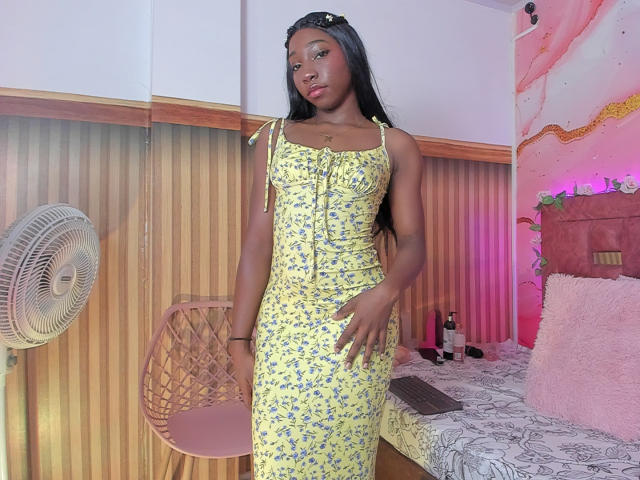 MelodyParis standing in a room, wearing a yellow floral dress, black hair, medium tits, dark skin, right hand on hip, left hand by side, wooden chair and bed behind.