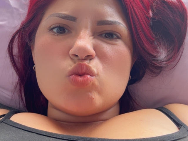 KatherineToro with red hair, pouty lips, and thick eyebrows, making a kissing face, showing her large tits and collarbone, wearing a black top.